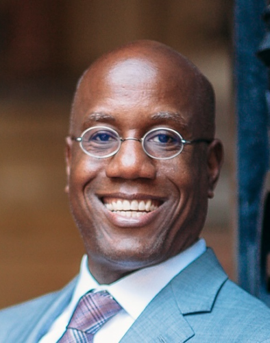 Bald man with glasses smiling, wearing a gray suit and patterned tie, posing for a photo.